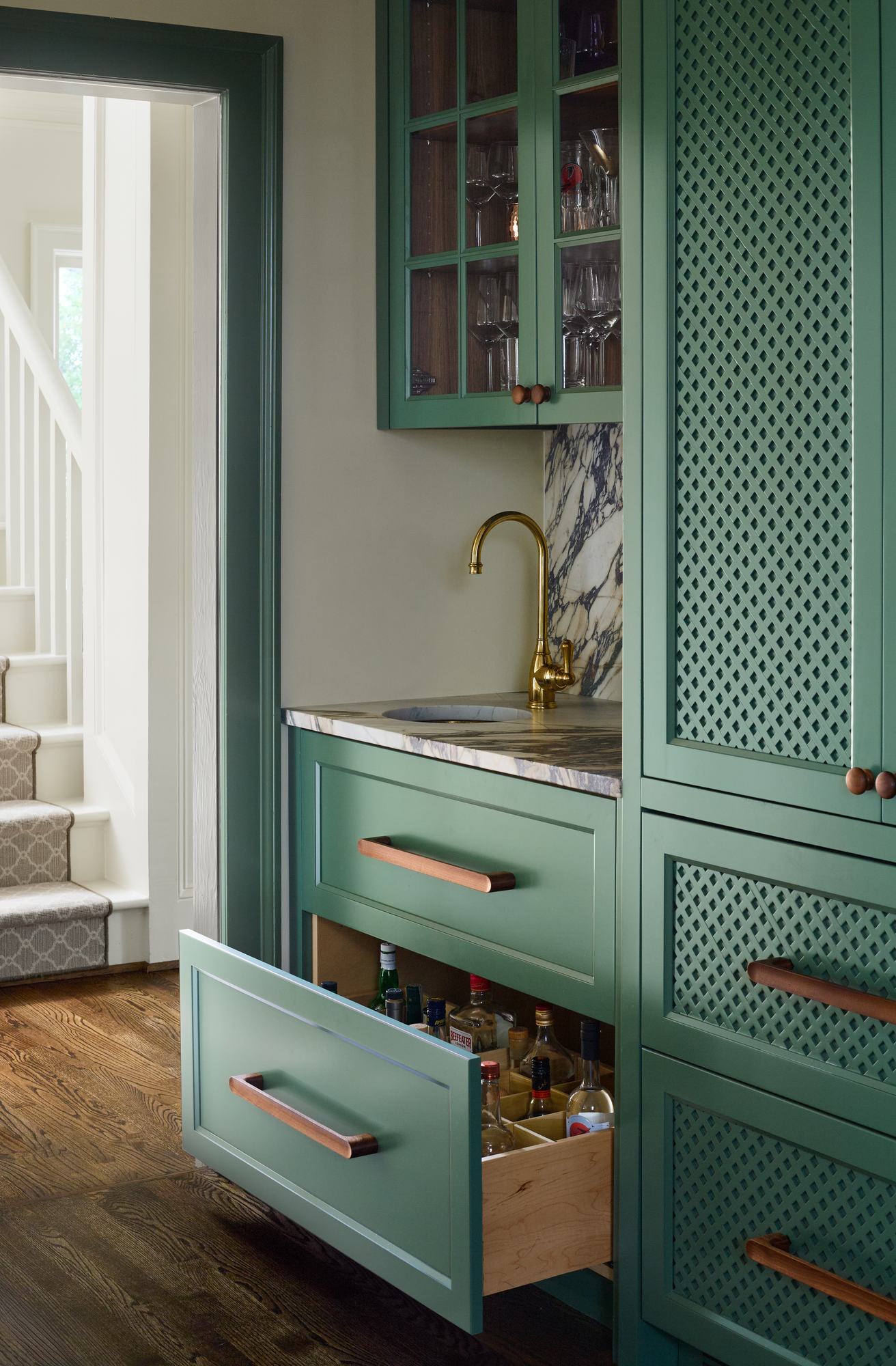 Open butler's pantry drawer with glassware and bar bottles behind green cabinetry